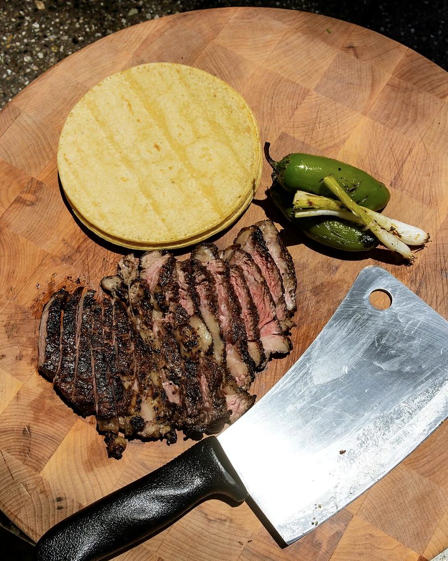 Sliced charred ribeye with corn tortillas, jalapeños and a cleaver on a wood board